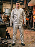 A man stands in a smoky industrial workshop wearing an Aluminized Kevlar long-sleeve shirt and pants set with a mottled, textured pattern, along with dark gloves and boots.