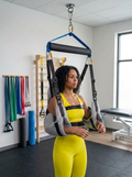 A woman in a bright yellow athletic outfit stands in a fitness studio using a hanging arm support harness with shoulder and elbow supports.