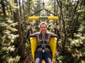 A woman smiling while riding a zip line suspended in a forest, seated in a yellow harness with a visible 'NewMeWindsCo.com' label on the bar above her.